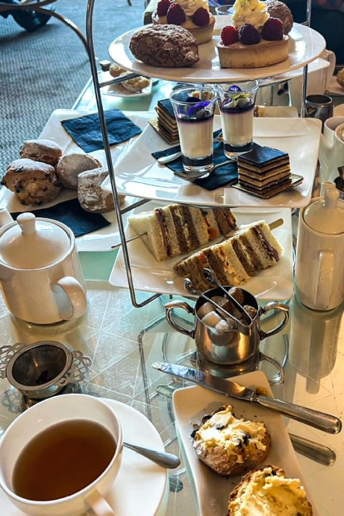 Tiered tea tray.  There are cakes, scones and finger sandwhiches with tea pot in the background