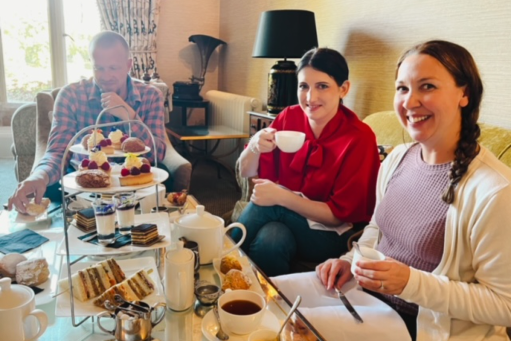 Friends sitting down having tea.  There is fancy sandwiches and pastries in front of them on a three tier plate. 