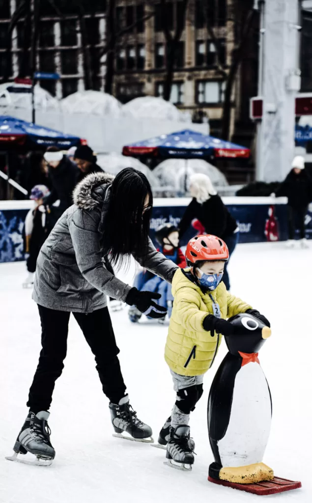 Small child with yellow coat and red helmet ice skating with his mom