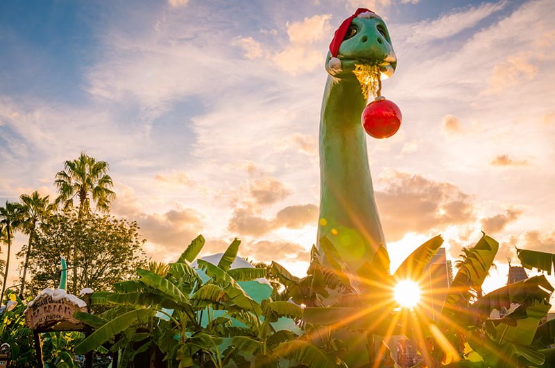 Dinosaur at Hollywood studio with a santa hat on and a christmas ball hanging from his mouth