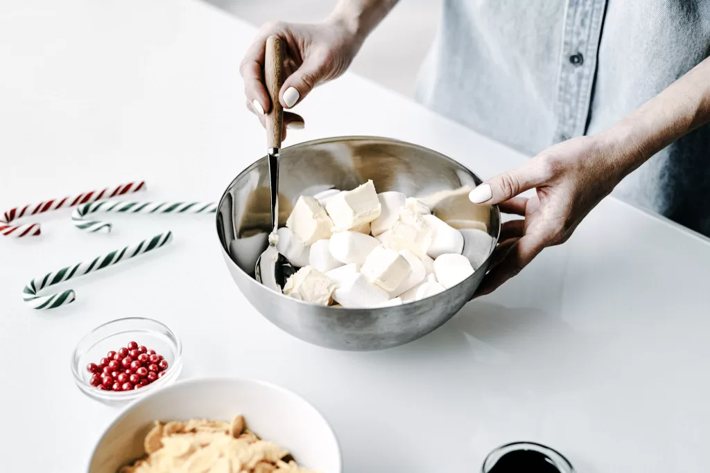 Preparing holiday treats.  There is a women;s hands mixing indrefients in a bowel with candy canes and red hots on the counter beside her. 