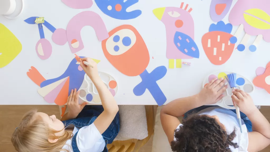 Activity table with white background and blue, red, yellow shapes.  Two children are sitting at table playing. 