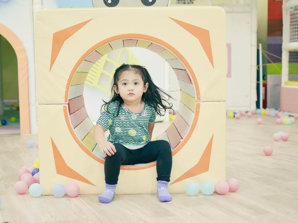 Small girl with black pigtails sitting on a colorful play tunnel at a playground.  Tunnel is orange and yellow