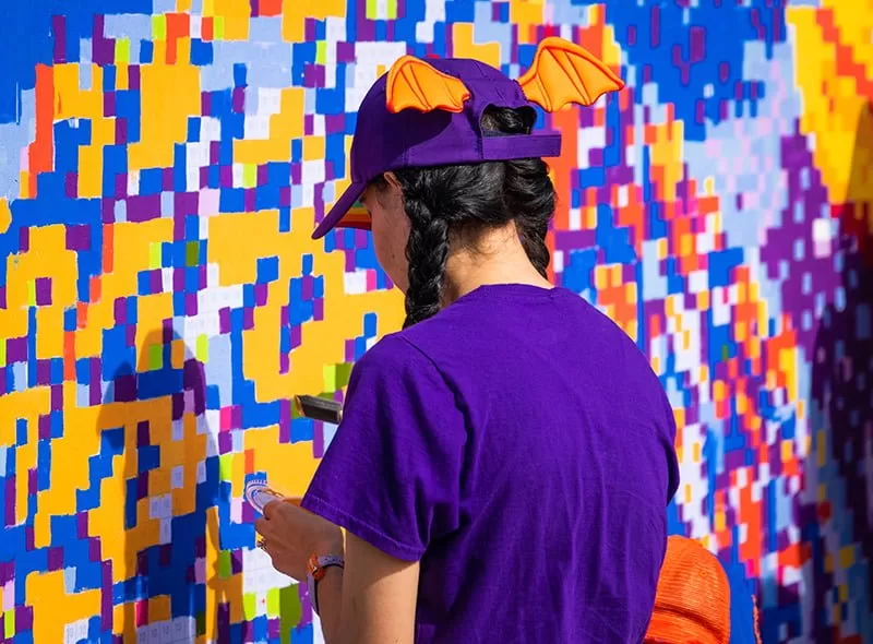 Girl in purple shirt standing in front of a mural painting it. 