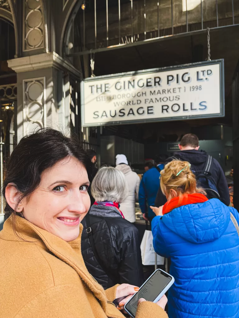 Women with brown hair and brown coat standing in ftont of a food stall called "the ginger pig"