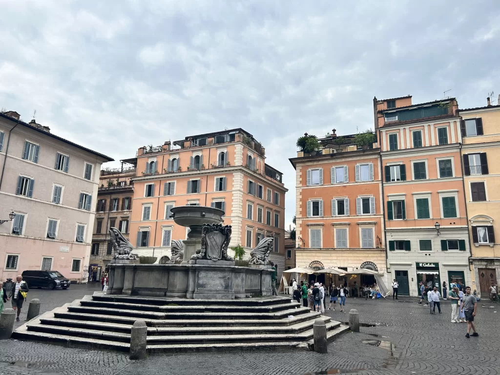 Large open square in Rome with old buildings surrounding the square.  There is a stone fountain in the middle. 