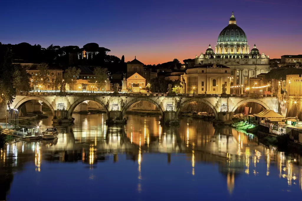 View of St Peter's basillicas large dome lit up at night with tiber river in the forground. 