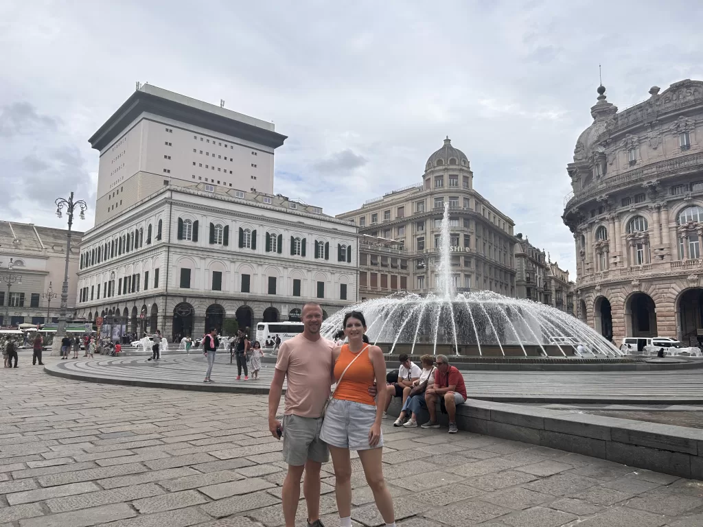 Man and woman stand in square, surrounded by tall white buildings in Genoa, Italy. 