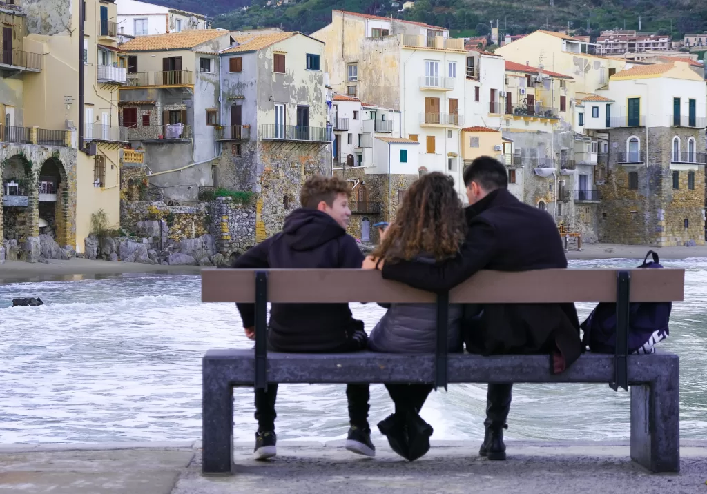 Mother, father, and teenage son sitting on a wooden bench in the middle of a square in Sicily