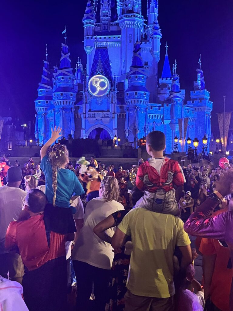 Two kids sitting on their parents shoulders in front of cinderellas castly at Mickey's not so scary halloween party
